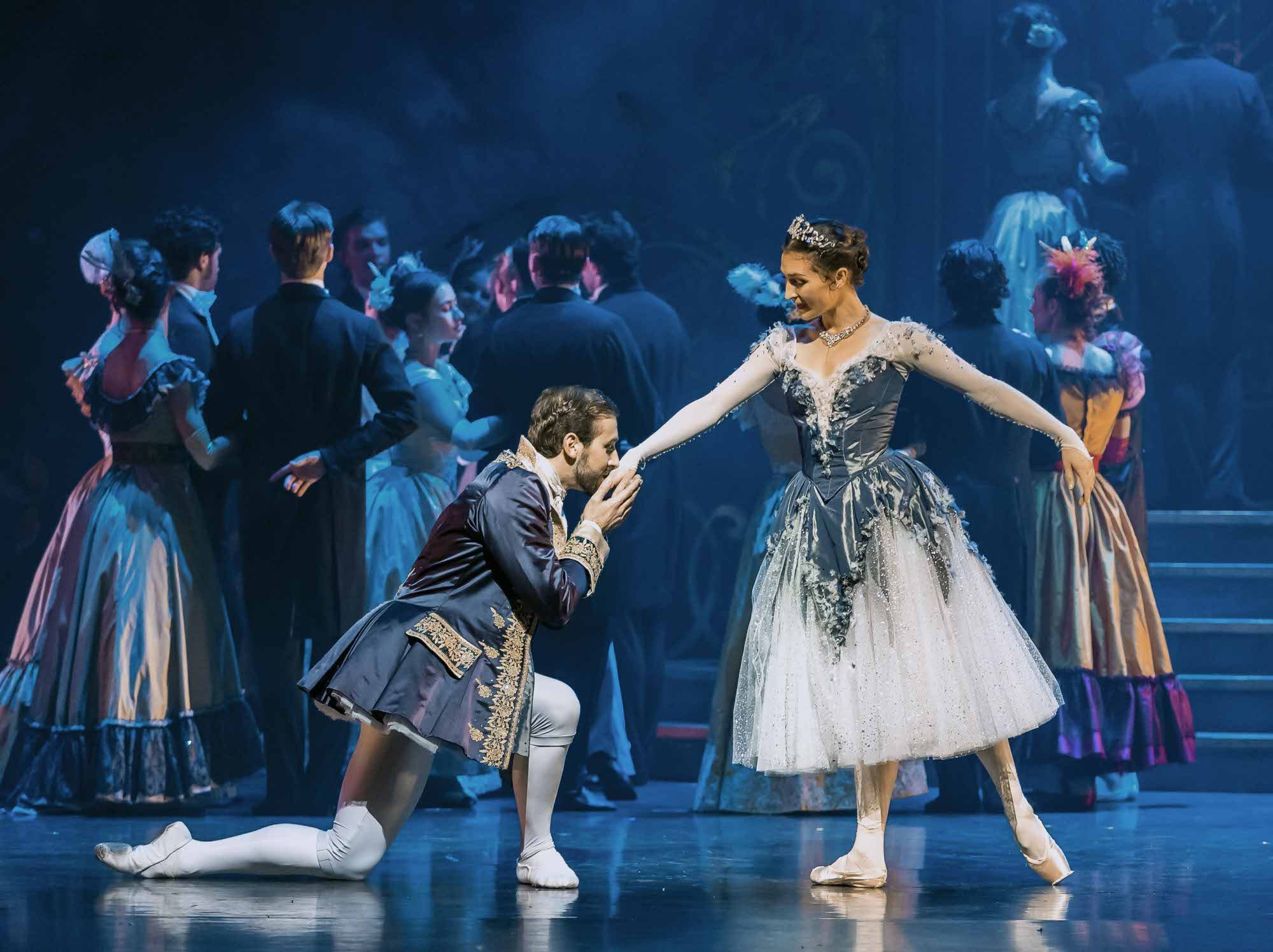 A man in a regal costume kneels and kisses the hand of a smiling woman in a sparkling gown and tiara, while other elegantly dressed people dance in the background on a dimly lit stage.